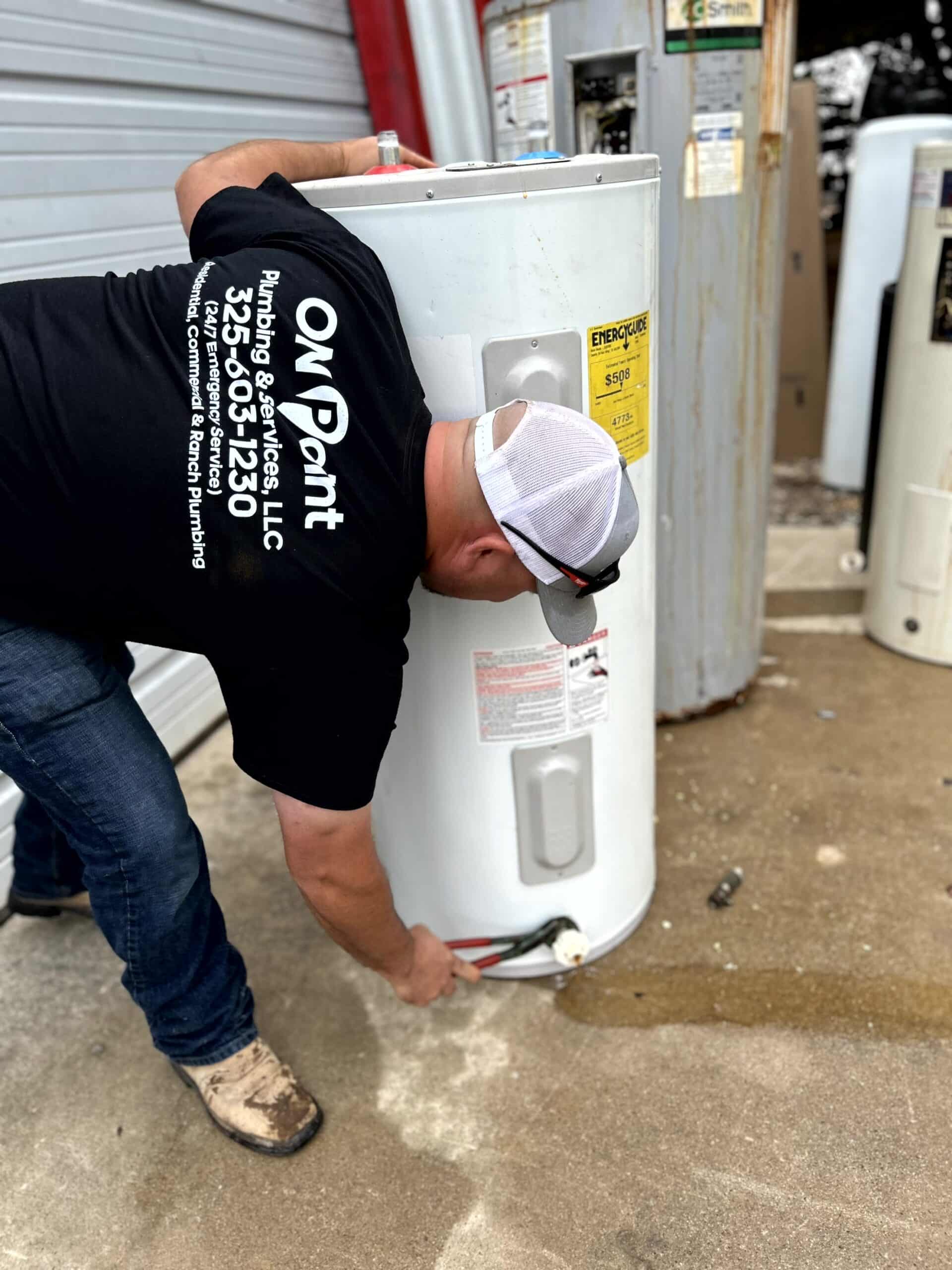 A plumber from On Point Plumbing working on a water heater installation, using a wrench to secure a connection. An old water heater stands nearby, showing signs of wear and rust.